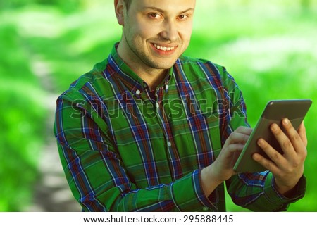 Gadget user concept. Portrait of funny hipster guy in green casual shirt reading e-book in park. White shiny smile. Bristle on face with healthy skin. Copy-space. Urban style. Outdoor shot