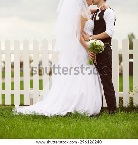 Happy married couple playing hugging and kissing each other. Bride holding wedding bouquet of beige flowers over white wooden fence and green lawn. Country vintage style. Outdoor shot