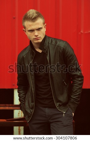 Stylish bully concept. Portrait of brutal young man with short hair wearing black jacket and posing over red urban background. Hipster style. Hands in pockets. Outdoor shot