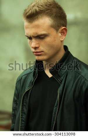 Stylish bully concept. Portrait of brutal young man with short wet hair wearing black jacket and posing over urban background. Hipster style. Close up. Outdoor shot