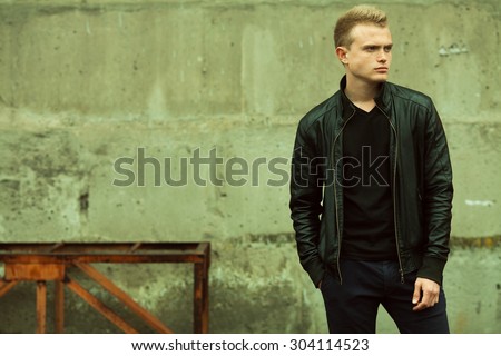 Bad boy concept. Portrait of brutal young man with short hair wearing black jacket, jeans and posing over urban background. Hand in pockets. Hipster style. Copy-space. Outdoor shot