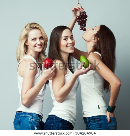 Raw, living food, veggie concept. Portrait of three happy young women wearing white sleeveless shirts, holding fruits over gray background. Casual clothing. Perfect skin, natural make-up. Studio shot