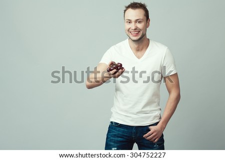 Raw, living food concept. Portrait of happy young man wearing white t-shirt, blue jeans, holding grape in hands over gray background. Casual clothing. Muscular body. Copy-space. Studio shot