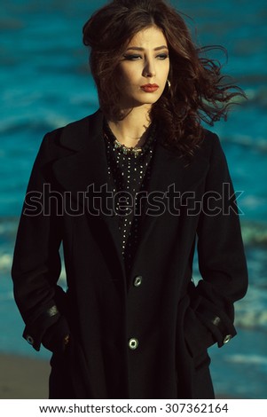 Bella donna concept. Beautiful brunette with long curly hair in black coat. Luxurious golden accessories: earrings, necklace. Perfect make-up. Street & Italian style. Windy weather. Outdoor shot