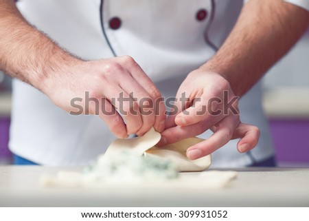 Vegetarian bakery concept. Chef cook in uniform putting filler of spinach and soft feta cheese on slice of puff pastry and making french chausson. Close up. Indoor shot