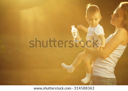 Happy family concept. Playing mother and little son in white casual sleeveless shirts. Toddler in white socks, diaper holding bottle of water. Rays of light at summer sunrise. Copy-space. Outdoor shot