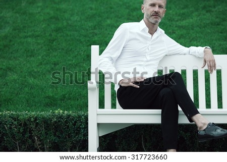 Men fashion concept. Portrait of mature charismatic man with blue eyes and beard wearing white shirt and black pants sitting on bench. Silver short hair. Iconic classic style. Copy-space. Outdoor shot