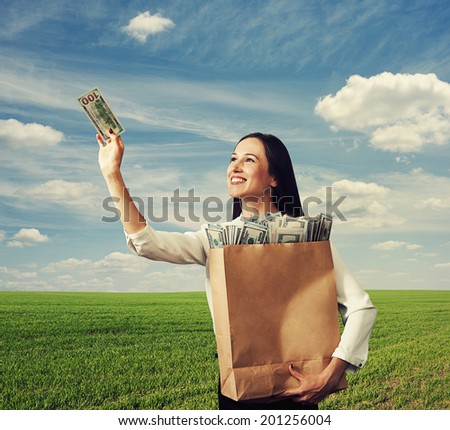 smiley successful woman holding paper bags with money and looking at one hundred dollars over blue sky and green field