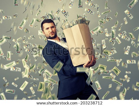 handsome successful businessman holding heavy paper bag with money under dollar\'s rain