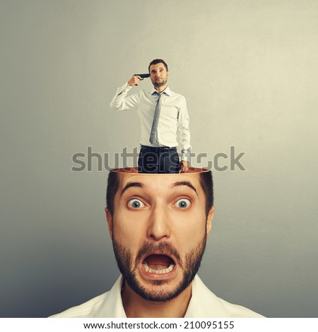 small sad man with gun standing in the head of shocked man. photo over grey background