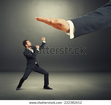 concept photo of conflict between subordinate and boss. angry young businessman showing fist and looking up at big palm of his boss. photo in the dark studio