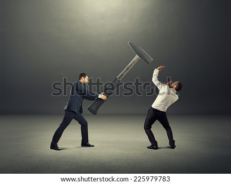 angry screaming businessman holding big hammer and hitting scared man. photo over dark background