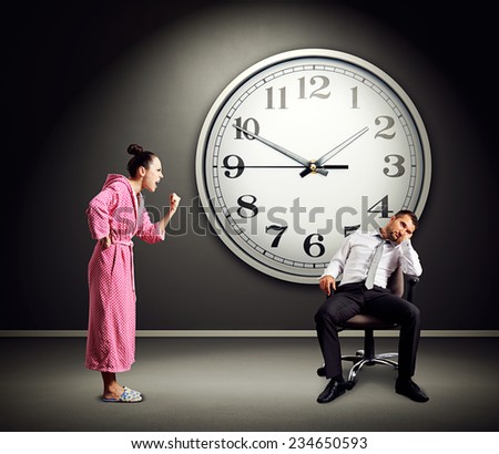 angry wife screaming at lazy husband. photo in dark room with big clock on the wall