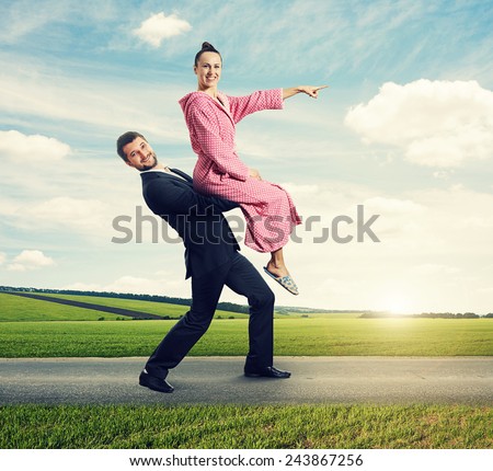 happy man walking on the road, carrying smiling woman. young woman sitting on man and pointing at the way