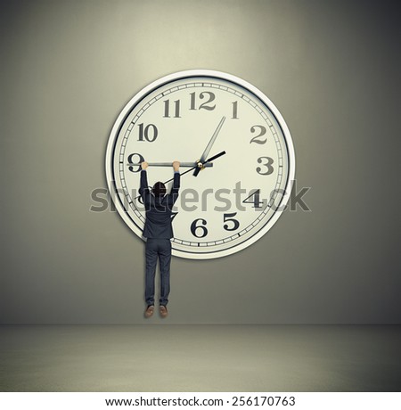 businessman hanging on big clock hand. big clock on grey wall