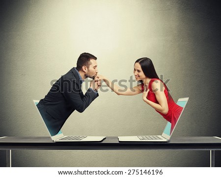 concept of love in social network. young man got out of the computer and kissing hand of beautiful young woman who got out of another computer over dark grey background