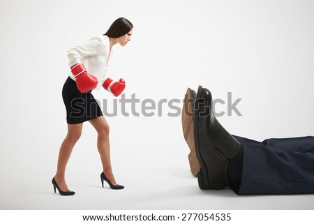 angry businesswoman in boxer gloves looking at big knocked out businessman over light grey background