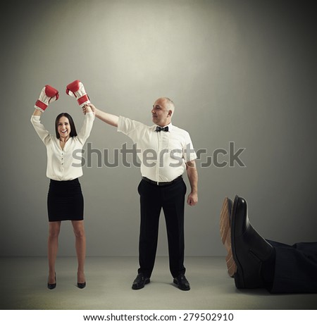smiley referee holding winner businesswoman in boxer gloves and looking at her, near lying big legs of knocked out businessman over dark grey background