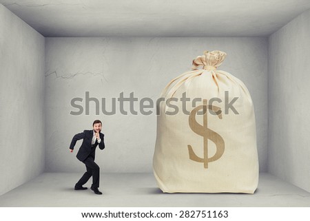 businessman in formal wear showing silent sign and walking on tiptoe to the big bag of money in grey room