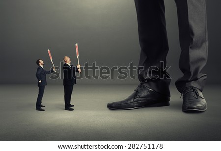 two small screaming businessmen holding placard and looking up at big legs over dark grey background