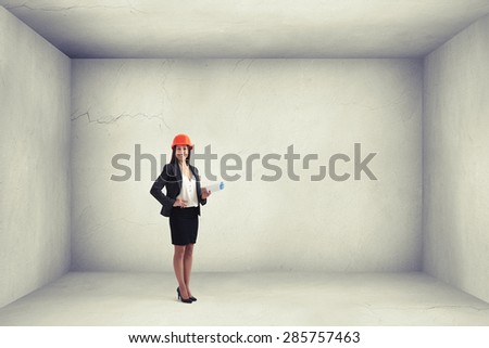 smiley woman architect in orange hardhat holding plan and looking at camera in empty concrete room with blank copyspace on the right