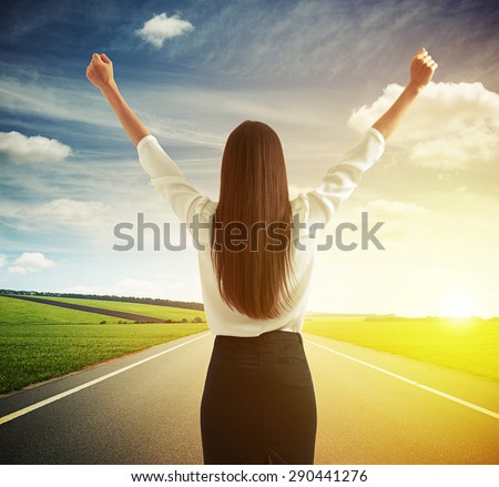 back view of happy woman raising hands up over road, green fields and sky