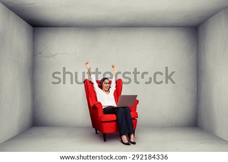 excited happy businesswoman with raised hands up sitting on the red chair and looking at laptop in grey empty concrete room