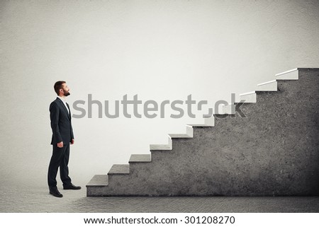 smiley businessman in formal wear standing near concrete stairs and looking at the top over light grey background