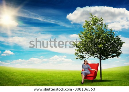 smiley young woman resting in red chair on green meadow near tree against blue sky