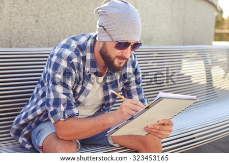 stylish bearded man sitting on bench and writing in notepad at outdoors