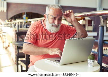 cheerful senior man raised his glasses and looking at laptop in cafe