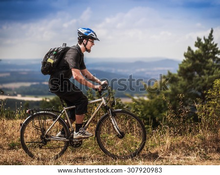 Biking in the Franconian Hills in Northern Bavaria. Caucasian Young man in black sports outfit with his Trekking bike on a rock, looking into the distance. Shot in High Summer August on an afternoon