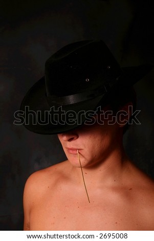Shirtless male model in Cowboy Hat chewing on a grass stem casting a shadow over his eyes.