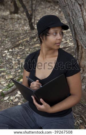 Young african-american woman sitting in forest, looking over her shoulder with notebook