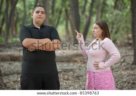 Angry wife pointing finger at her overweight husband, he looks a little insolent. They are outside, in a forested area.