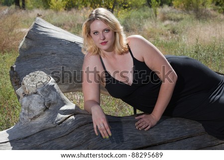 Beautiful full figured blonde woman wearing black dress resting on a log. She is wearing cowboy boots, and it is autumn.