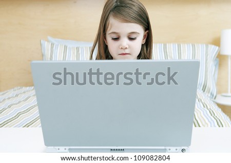 A little girl sitting at the foot of her bed behind a large laptop screen with only her head visible over the top.