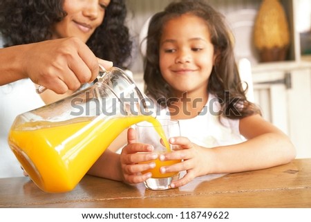 Mother pouring fresh orange juice from a jug for her daughter seated at a wooden table in the kitchen