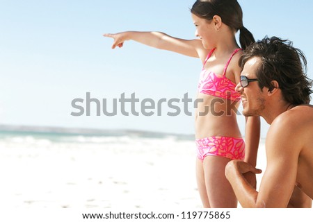 A young daughter points into the distance showing something to her father who is kneeling at her sideat beach