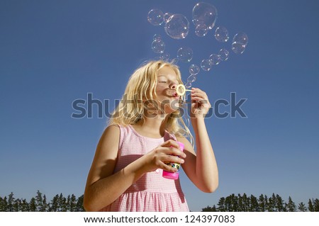 Pretty young blonde girl in a pink frock standing against a blue sky blowing bubbles