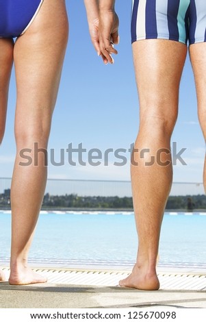 Man and woman standing side by side alongside a pool in bathing costumes cropped so that only one leg of each is visible
