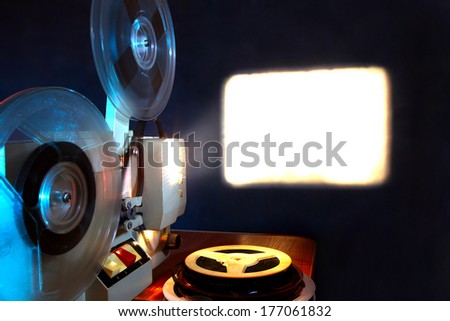 Old 8mm film projector showing the film in dusk onto a wall beside a stack of film reels