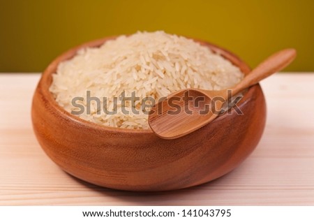 Uncooked rice in bamboo bowl with spoon on a wooden surface over dark yellow background