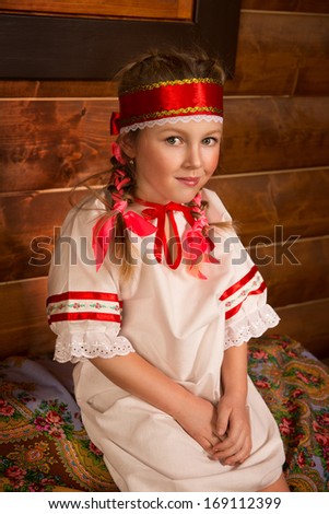 Girl in Russian national sundress. Folk dress.Cute little blonde girl in the hut. Sits on a table and smiling