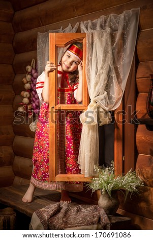 Closeup portrait of adorable blonde little girl in a scarf in russian national folk dress sits in a wooden hut stand near the window. Traditional dress on cute little girl who smile look at the camera
