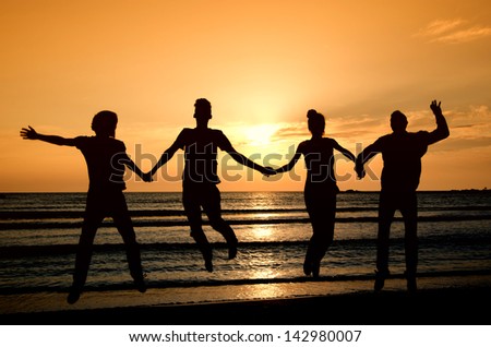 Group of happy people parting on the beach at sunrise, all holding hands and jumping of joy