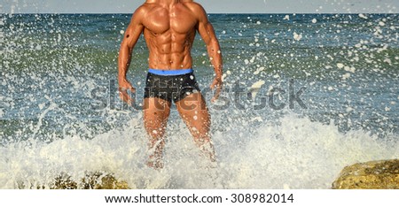 Strong bodybuilder with six pack.Fitness trainer with perfect abs, shoulders,biceps, triceps,chest, flexing his muscles on the beach with sea waves on the background, training in vacation