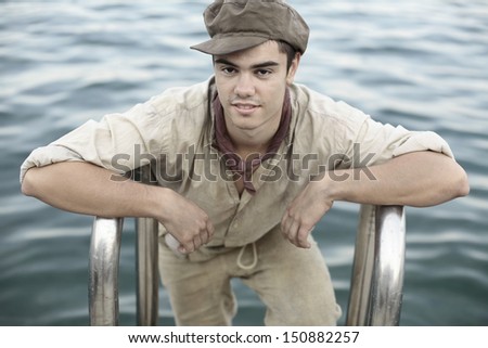 portrait of a boy in old retro clothes and cap standing by the water looking poor and dirty, an apprentice