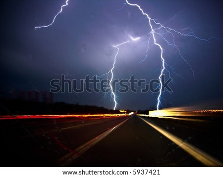 Two forks of bluish-white lightning strike on either side of a dark highway at night, materialized by the streaks of taillights and headlights