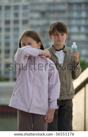 Two children stand in an urban setting, one sneezing into their elbow, the other holding a bottle of hand cleanser.